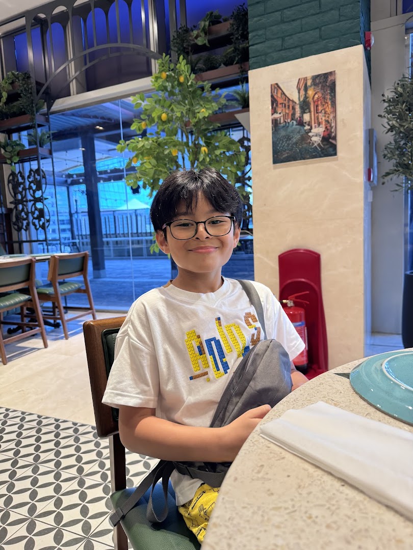 A young boy smiles while seated at a table in a bright, plant-decorated restaurant interior.