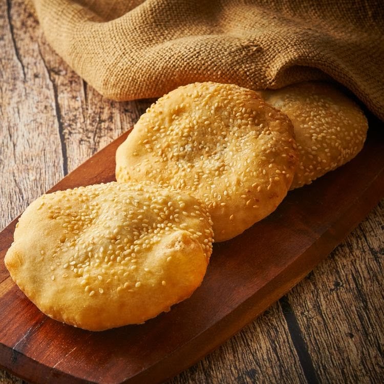 Three golden sesame flatbreads on a wooden board, with a burlap cloth in the background.