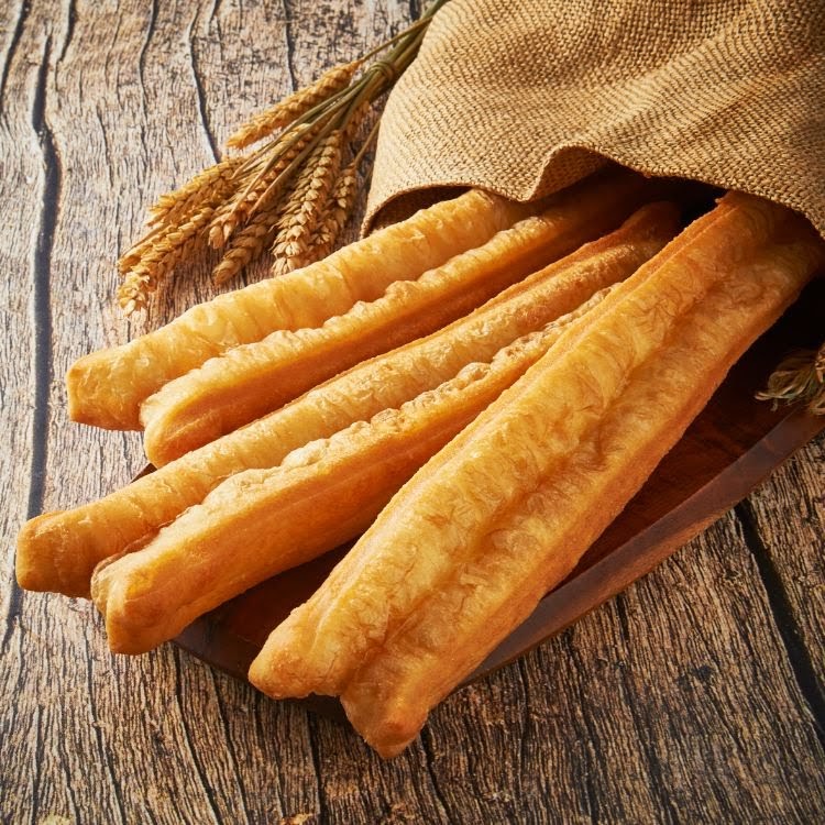 Golden fried dough sticks (youtiao) on a wooden board with wheat stalks and burlap.