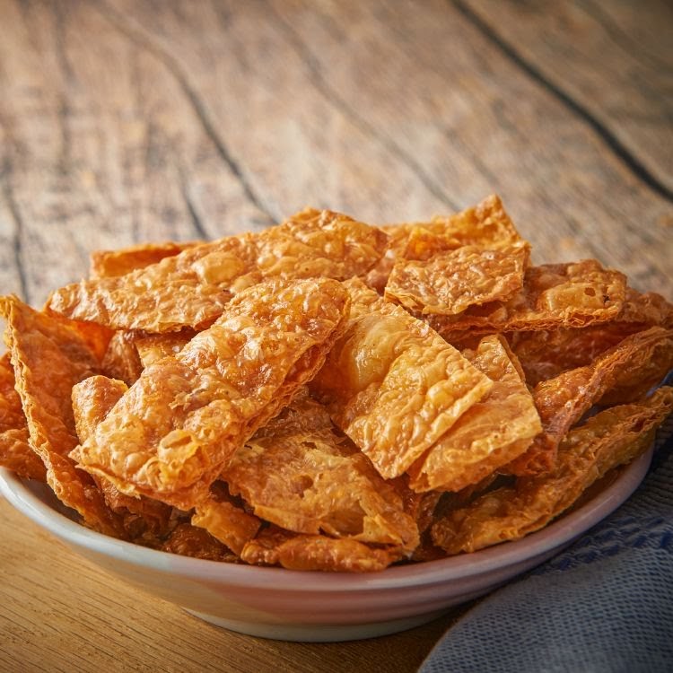 A bowl filled with golden-brown, crispy fried beancurd skin on a rustic wooden table.