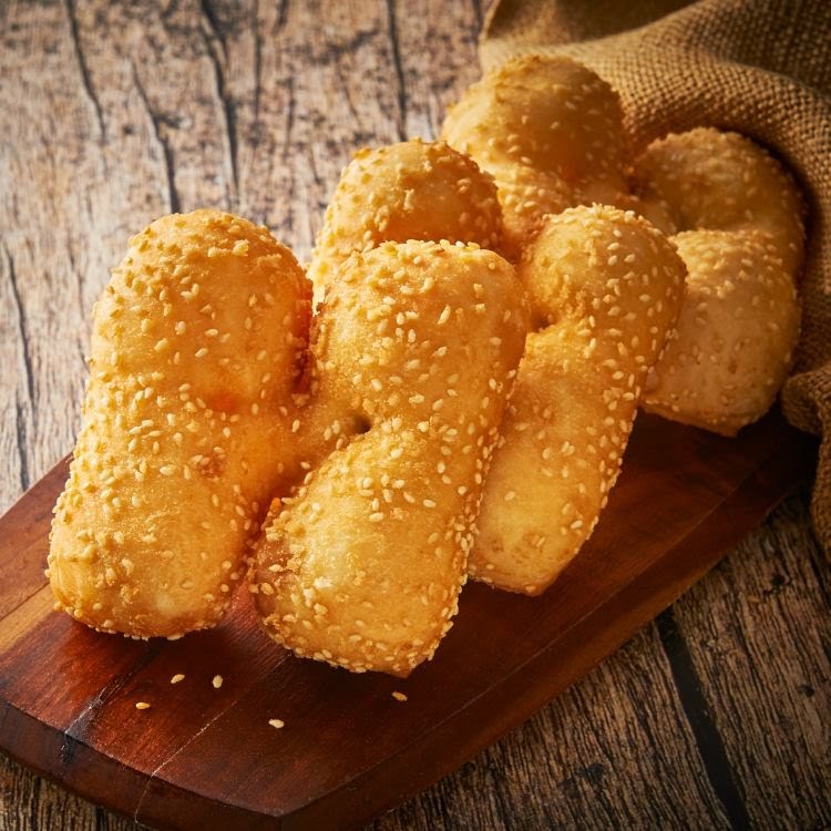 Golden fried dough sticks coated with sesame seeds, presented on a wooden board.