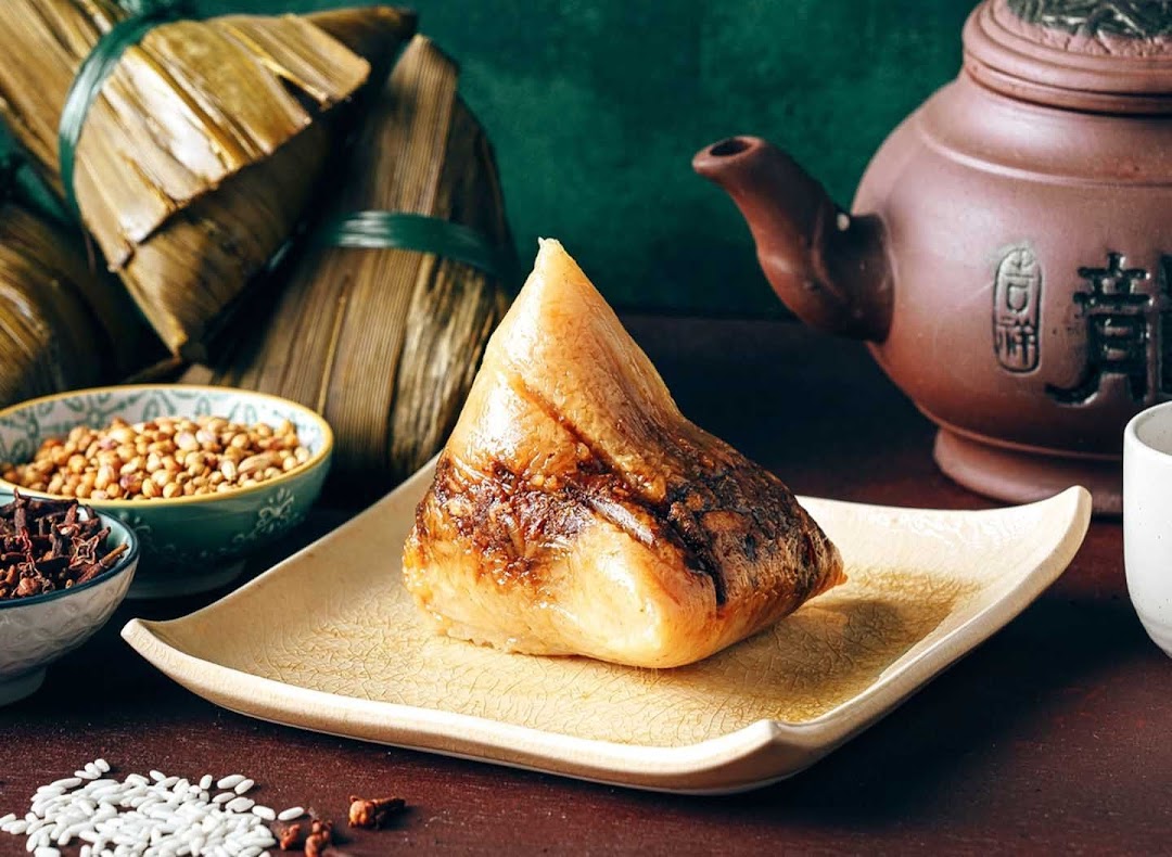 A delicious, unwrapped rice dumpling (zongzi) on a plate, with wrapped dumplings and tea in the background.