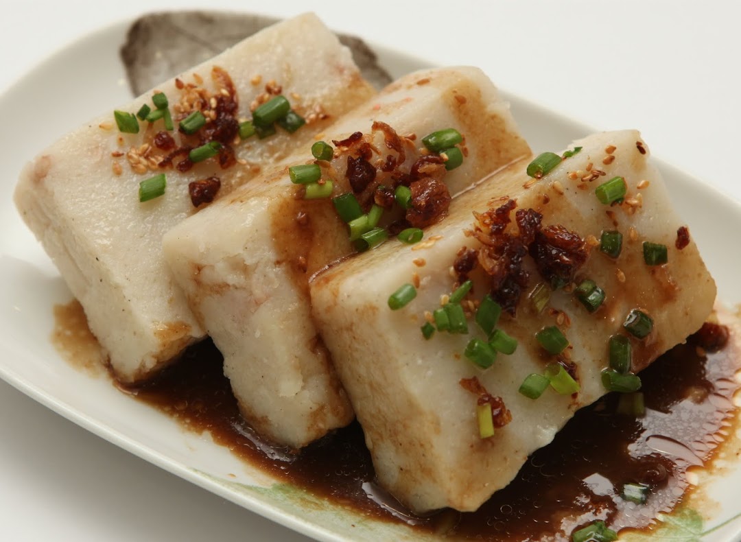 Three pieces of steamed radish cake with savory sauce and green onions on a white plate.