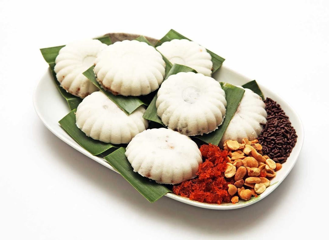 A plate of white, fluted cakes on banana leaves with peanuts, chili, and chocolate sprinkles.