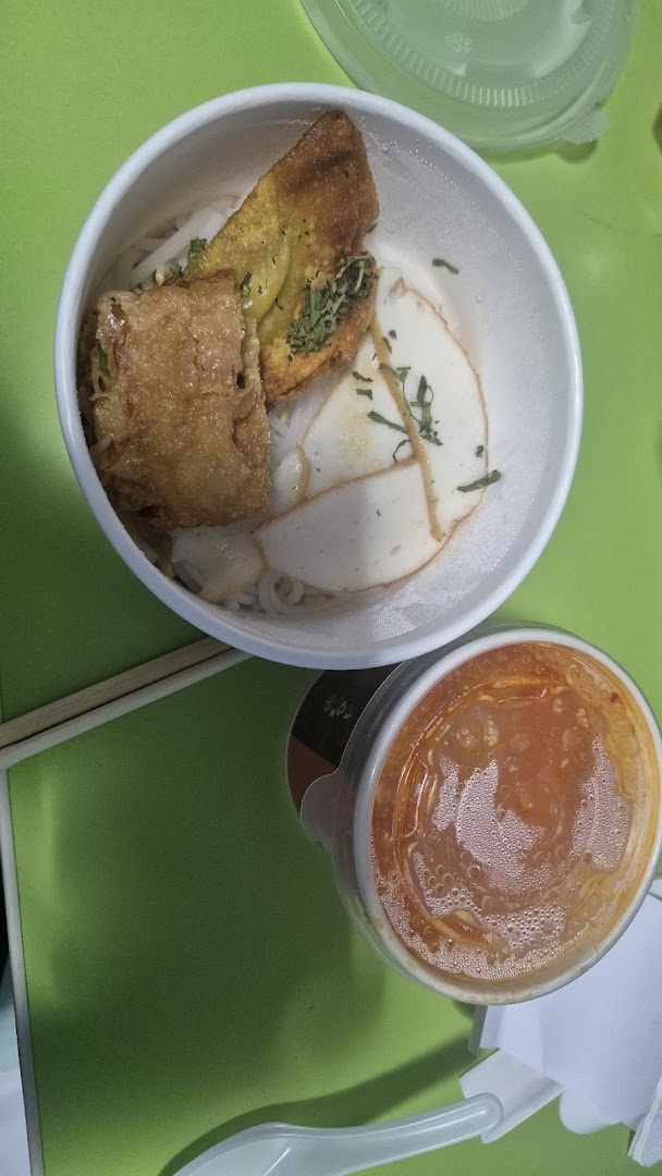 A close-up of a delicious noodle bowl with fried toppings and fish cakes, alongside a cup of sauce.