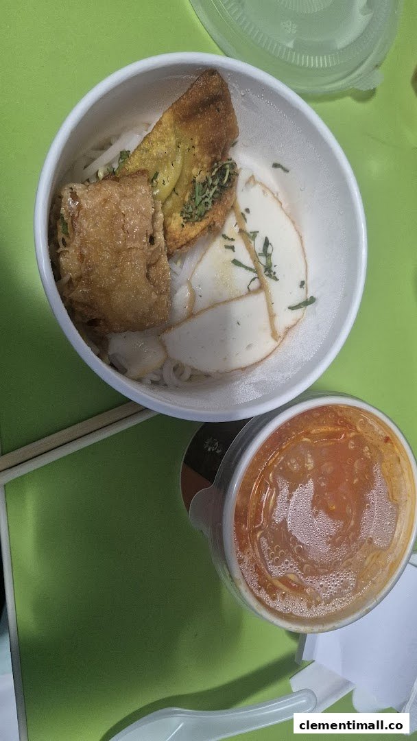 A close-up of a delicious noodle bowl with fried toppings and fish cakes, alongside a cup of sauce.