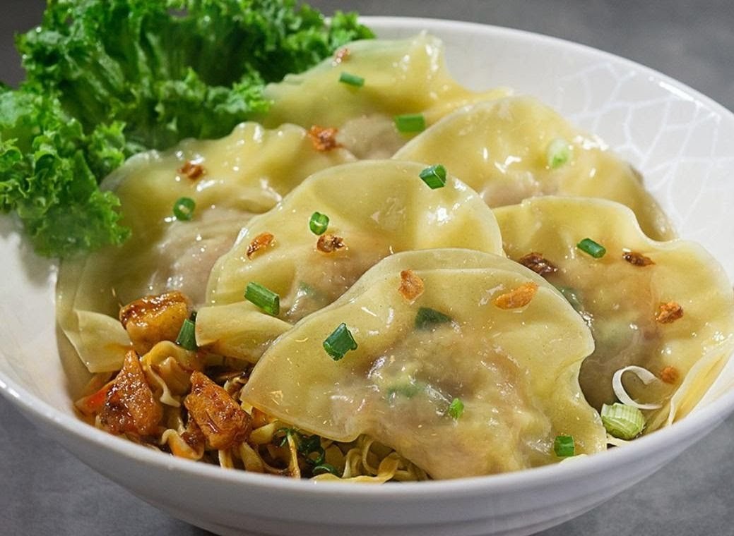 A close-up of delicious dumplings and noodles in a white bowl, garnished with greens and fried garlic.