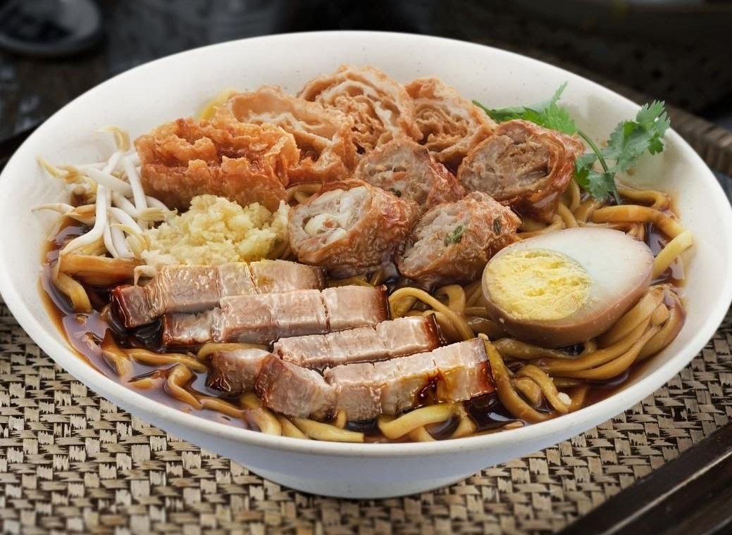 A close-up of a delicious bowl of noodles with sliced meat, fried items, bean sprouts, and a boiled egg.