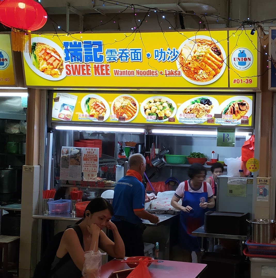 A bustling hawker stall named Swee Kee, specializing in Wanton Noodles and Laksa, with visible menu items and prices.