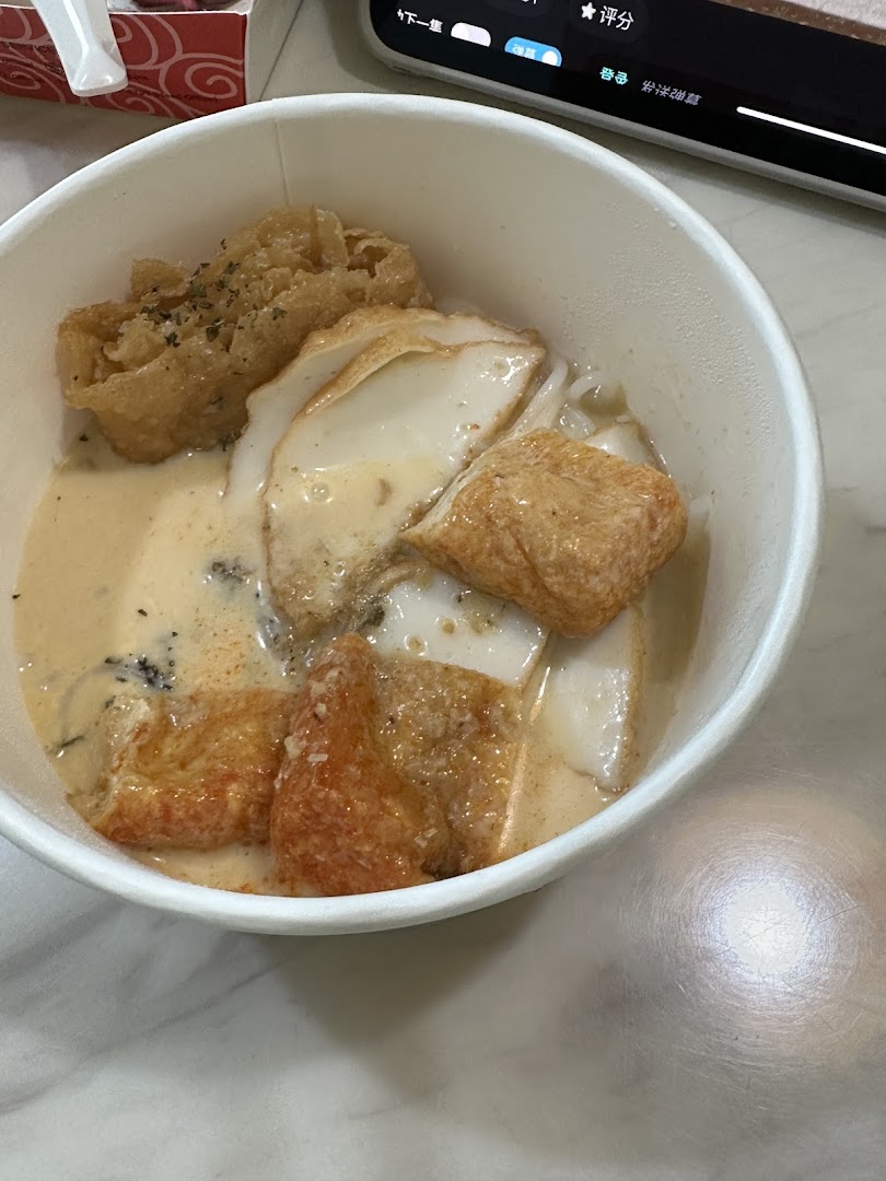 A close-up of a creamy noodle dish with various fried toppings in a white takeaway bowl.