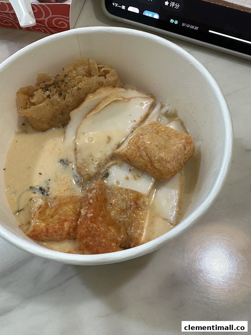A close-up of a creamy noodle dish with various fried toppings in a white takeaway bowl.