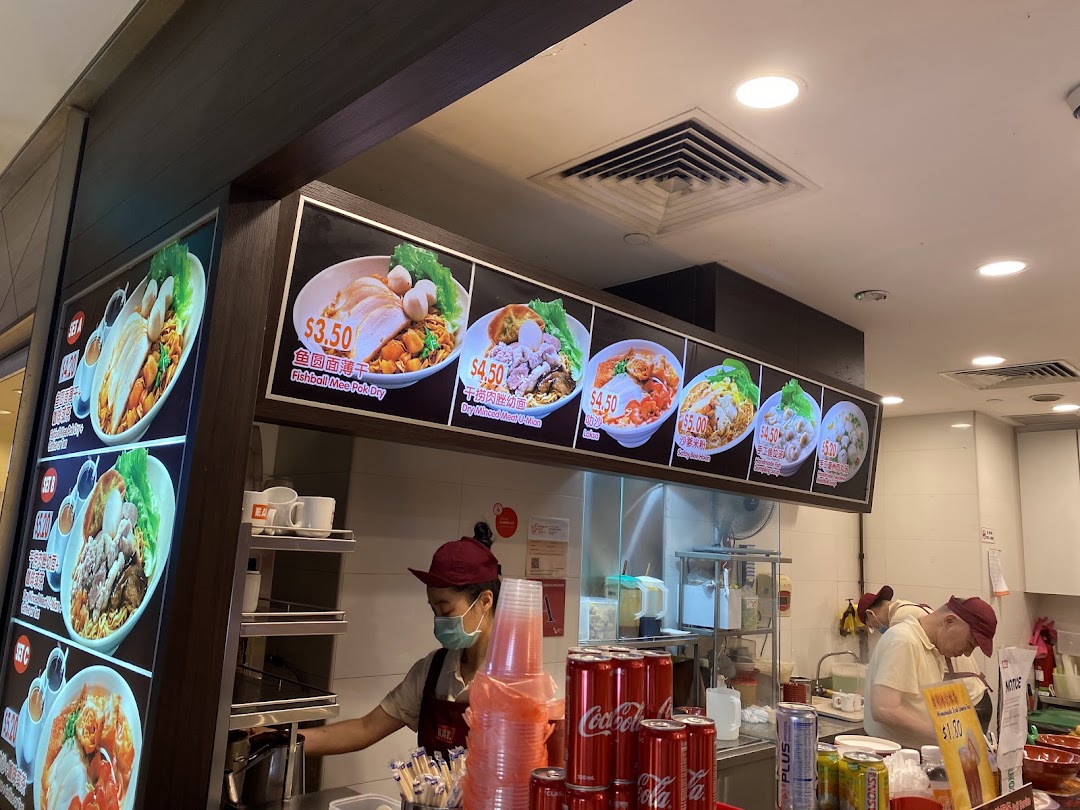 A food stall featuring large menu boards displaying various noodle dishes and prices, with staff preparing food.