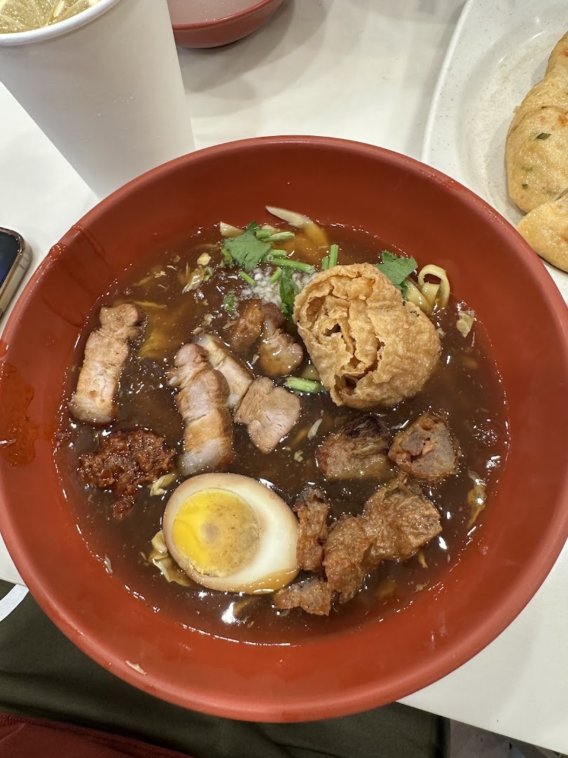 A close-up of a rich, dark noodle soup with pork, egg, and crispy toppings in a red bowl.