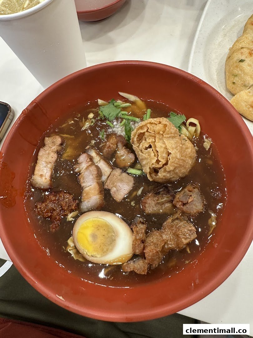 A close-up of a rich, dark noodle soup with pork, egg, and crispy toppings in a red bowl.