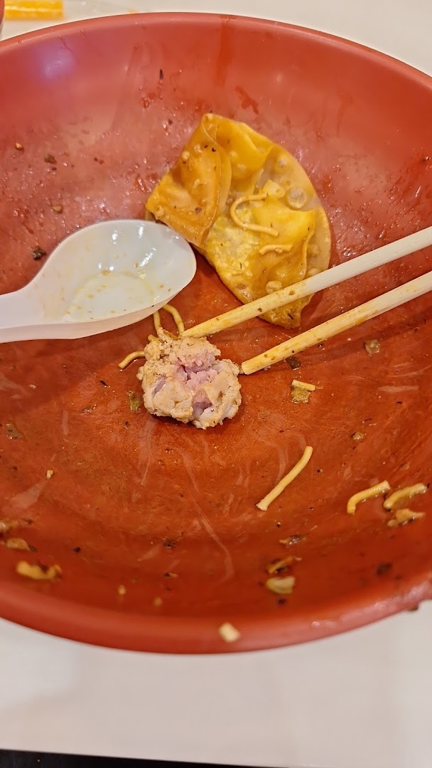 A red bowl with leftover food, including a dumpling, meat, noodles, a spoon, and chopsticks.