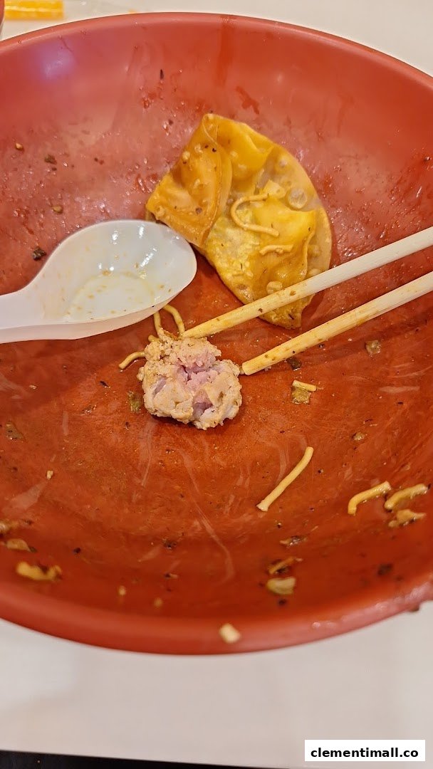 A red bowl with leftover food, including a dumpling, meat, noodles, a spoon, and chopsticks.