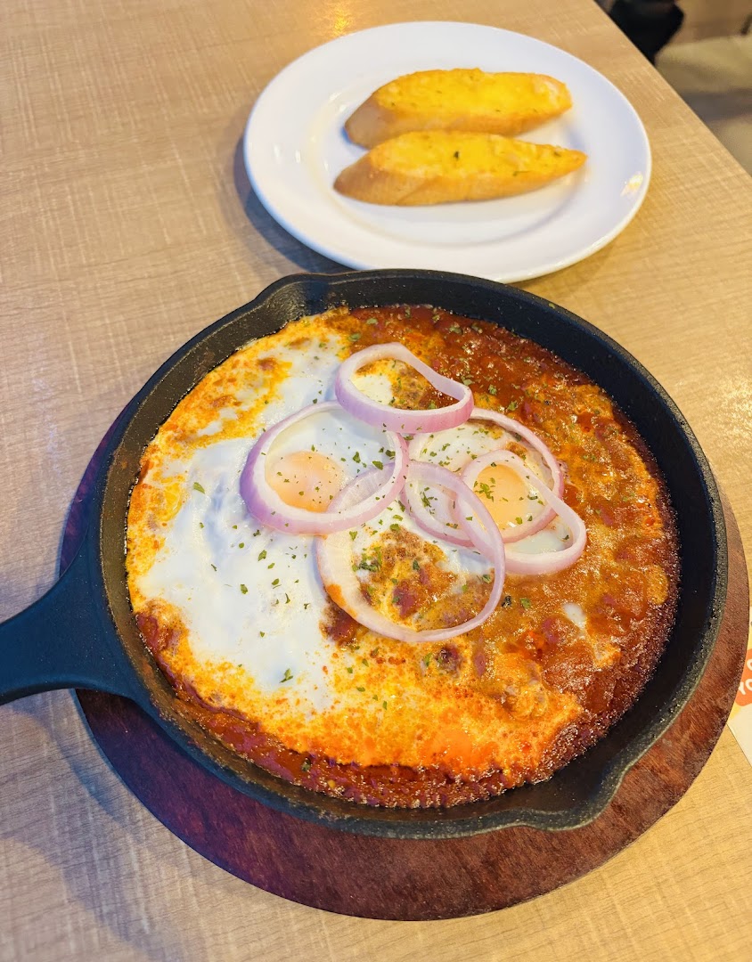 A sizzling skillet dish with baked eggs, tomato sauce, and red onions, served with garlic bread.
