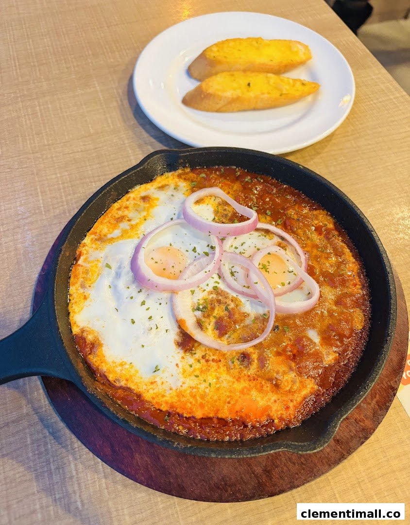 A sizzling skillet dish with baked eggs, tomato sauce, and red onions, served with garlic bread.