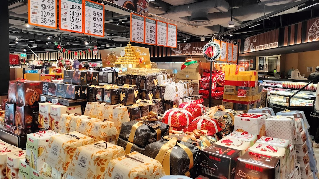 A supermarket aisle showcasing a wide array of festive cakes, panettone, and gourmet gift sets, with overhead price signs.
