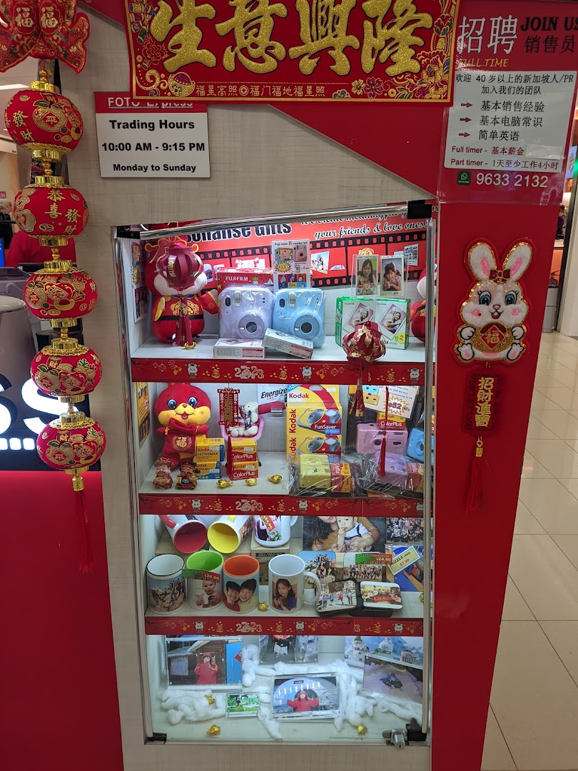 A retail display case featuring instant cameras, photo films, personalized gifts, and photo printing services, with shop hours.