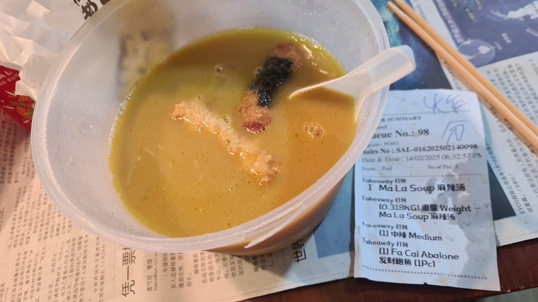 A close-up of a takeaway bowl of Ma La Soup with ingredients, a spoon, chopsticks, and a receipt.