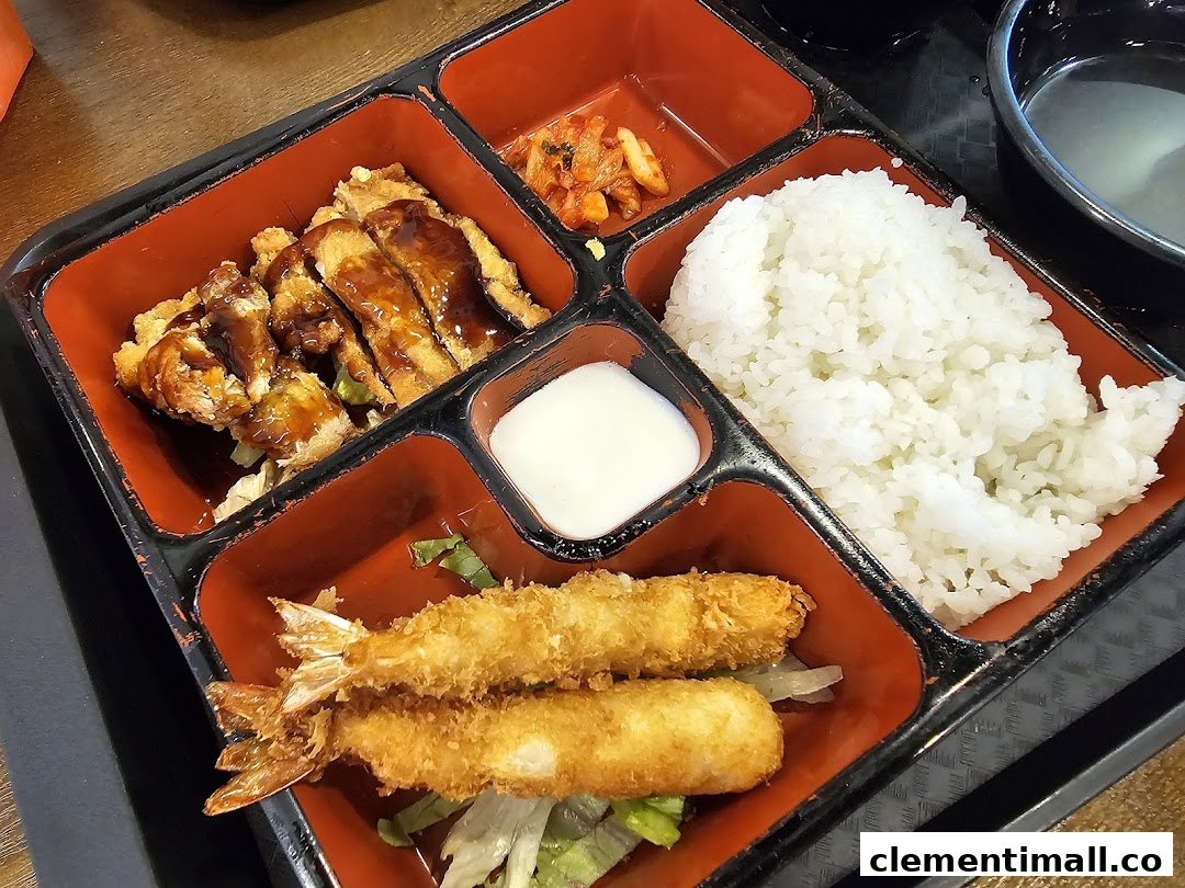 A vibrant bento box featuring crispy fried chicken, shrimp tempura, rice, kimchi, and a dipping sauce.