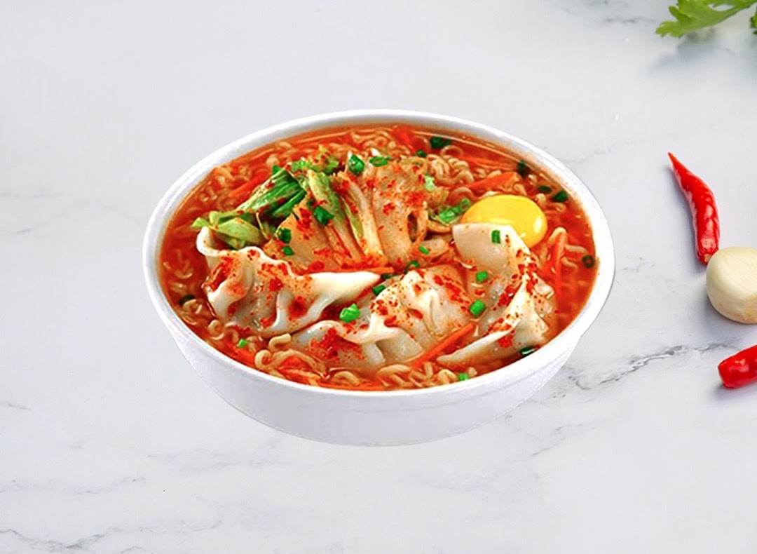 A vibrant bowl of Korean ramen with kimchi, dumplings, and an egg yolk, garnished with chili flakes.