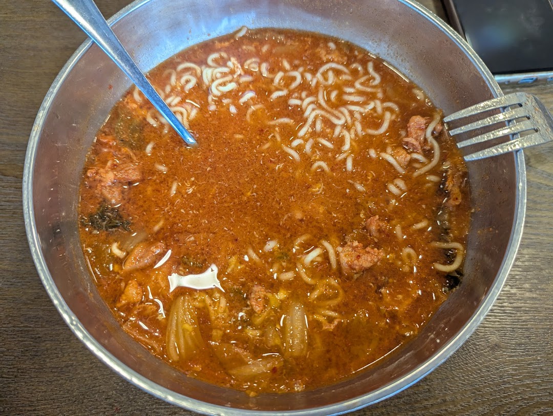 A close-up view of a vibrant red Korean ramen or stew in a metal bowl with noodles and ingredients.