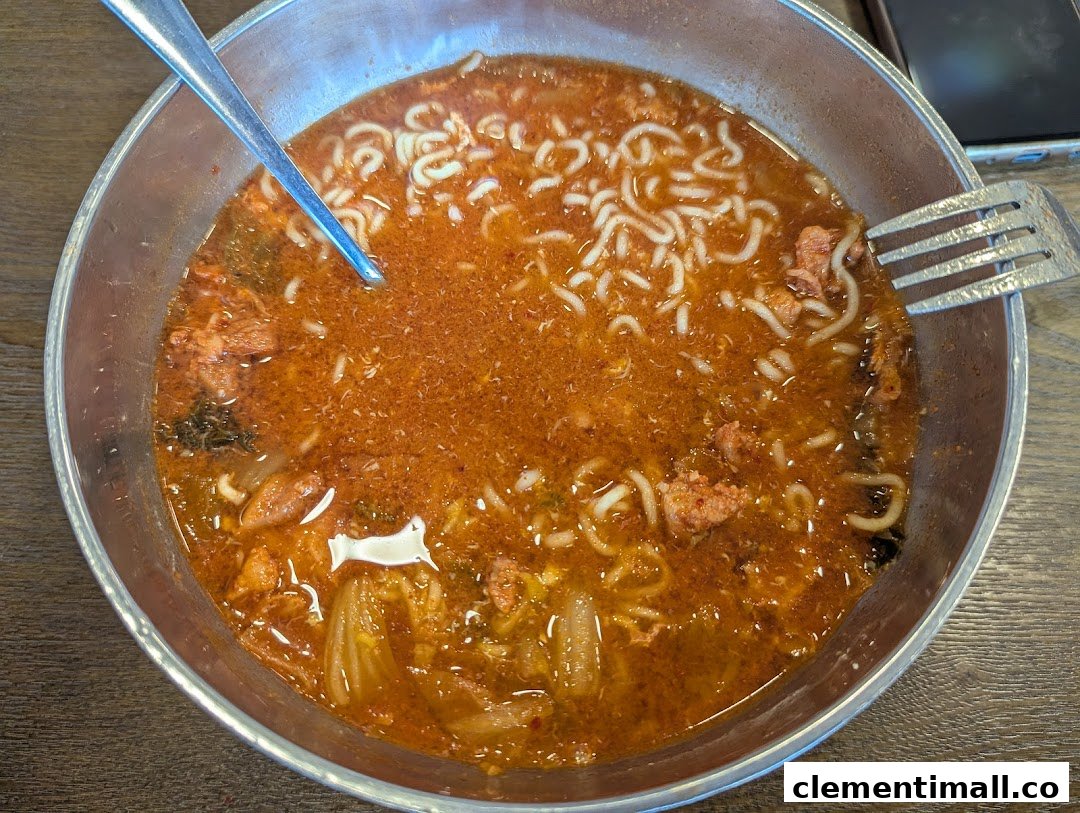 A close-up view of a vibrant red Korean ramen or stew in a metal bowl with noodles and ingredients.
