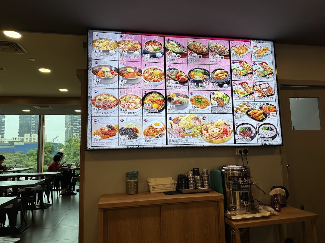 A brightly lit digital menu board displaying various Korean dishes with prices and images inside a restaurant.