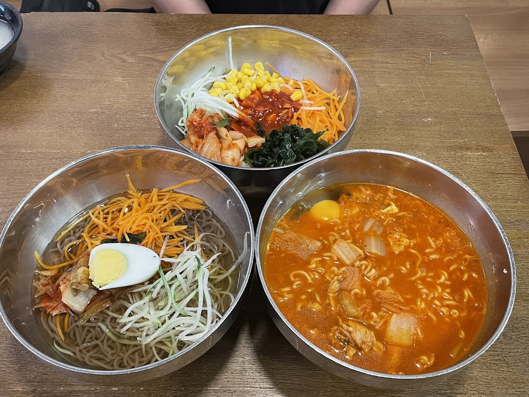 Three vibrant bowls of Korean dishes, including cold noodles, spicy ramen, and bibimbap, served on a wooden table.