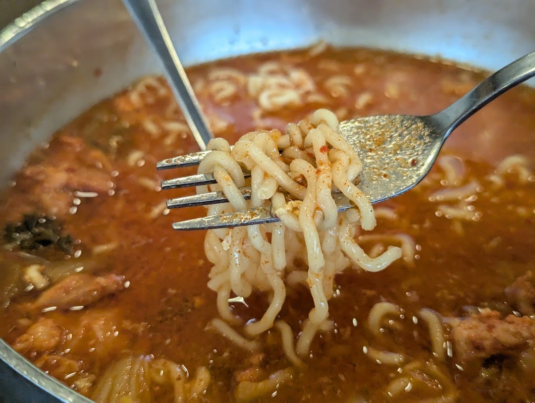 A close-up of a fork lifting spicy noodles from a steaming bowl of Korean ramen.