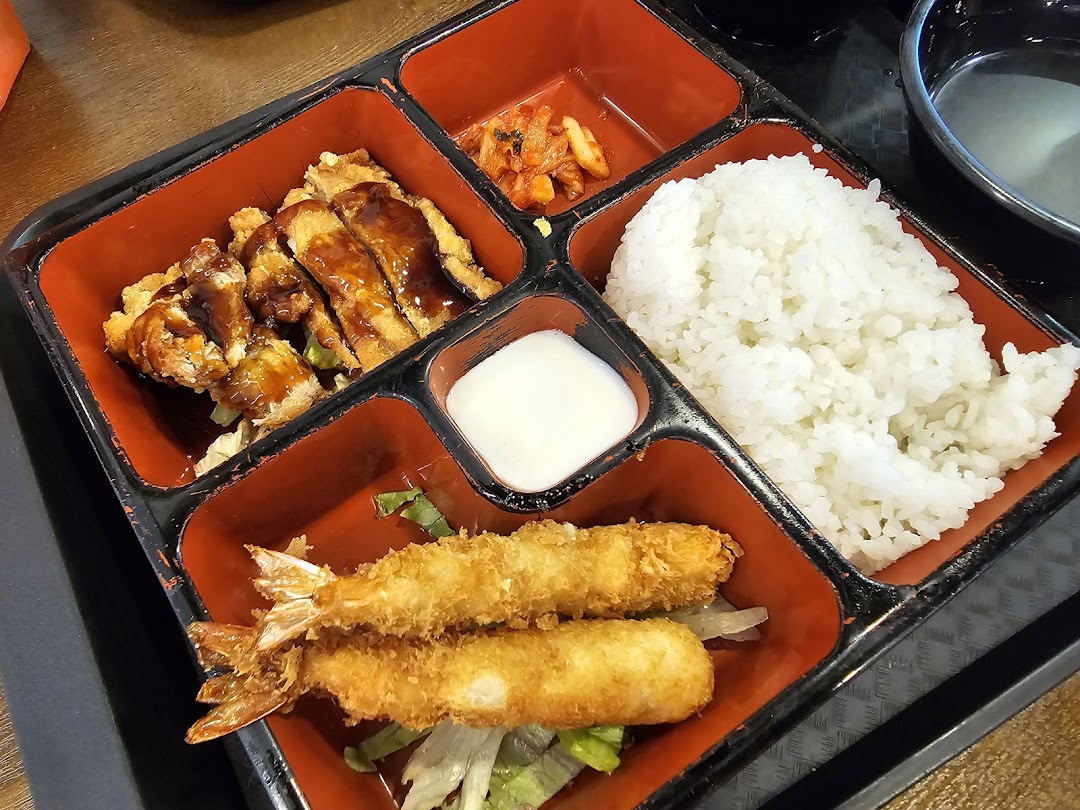 A vibrant bento box featuring crispy fried chicken, shrimp tempura, rice, kimchi, and a dipping sauce.