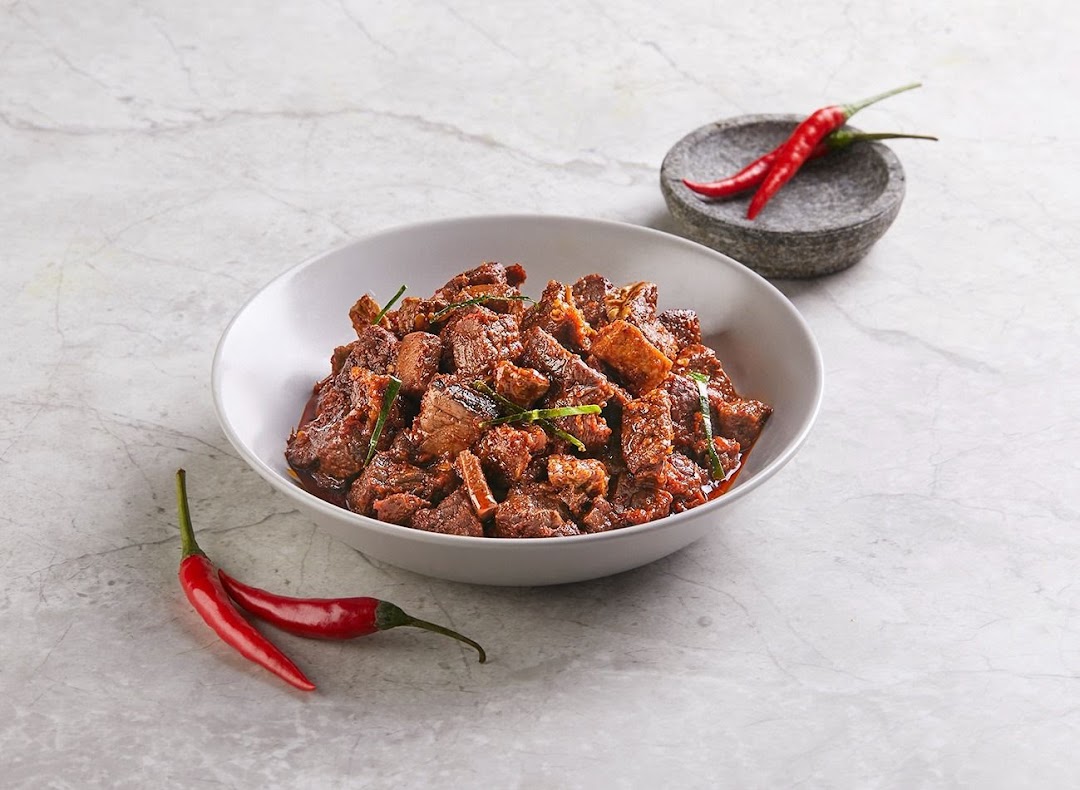 A bowl of spicy, rich meat stew, possibly beef rendang, garnished with green herbs and red chilies.