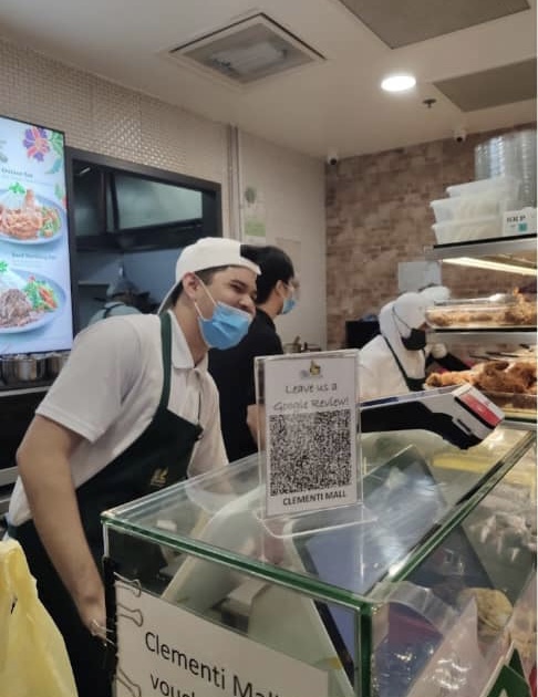 Friendly staff at Hjh Maimunah Mini serving customers, with a digital menu board in the background.