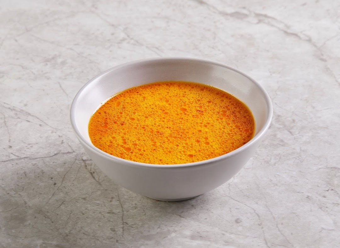 A white bowl filled with vibrant orange soup, possibly curry or broth, on a grey marbled surface.
