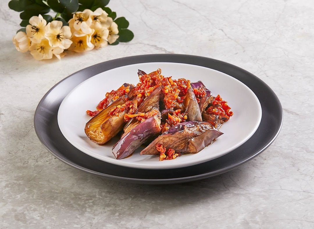 A plate of cooked eggplant topped with a vibrant red chili sambal, presented on a white and dark grey plate.