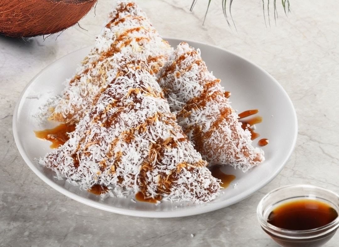 Three pyramid-shaped pastries covered in shredded coconut and drizzled with a sweet brown sauce, served on a white plate.