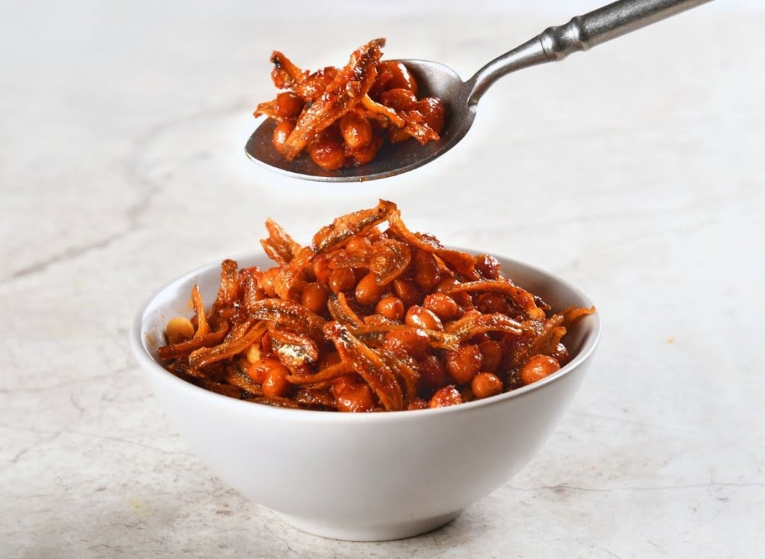A white bowl filled with crispy anchovies and peanuts in a sweet and spicy sauce, with a spoonful lifted above.