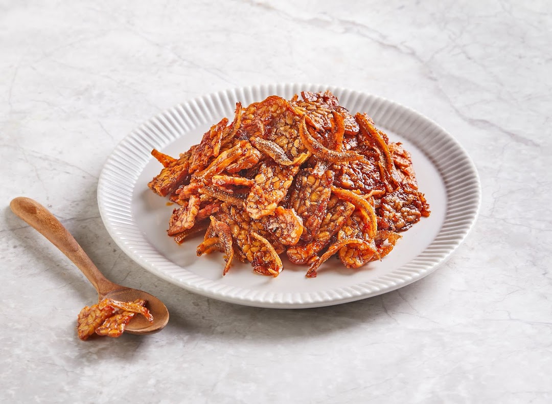 A plate of crispy, glazed tempeh and anchovies with a wooden spoon on a light marble surface.