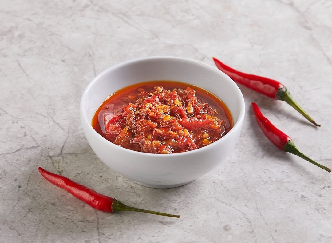 A white bowl filled with vibrant red chili sauce, surrounded by fresh red chilies on a light background.