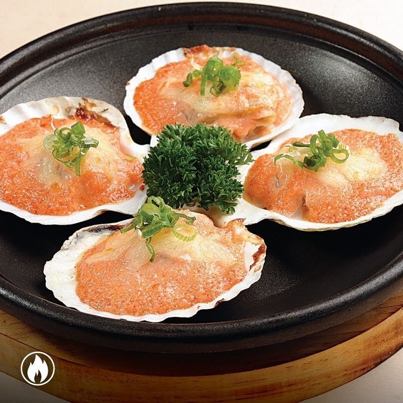 Four baked scallops in shells, topped with cheese and spring onions, served on a dark plate.