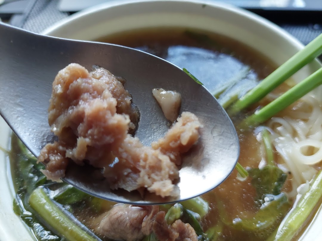 A close-up of a spoon lifting meat from a bowl of Thai noodle soup with noodles and greens.