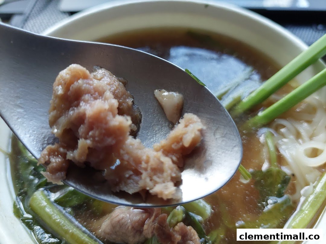 A close-up of a spoon lifting meat from a bowl of Thai noodle soup with noodles and greens.