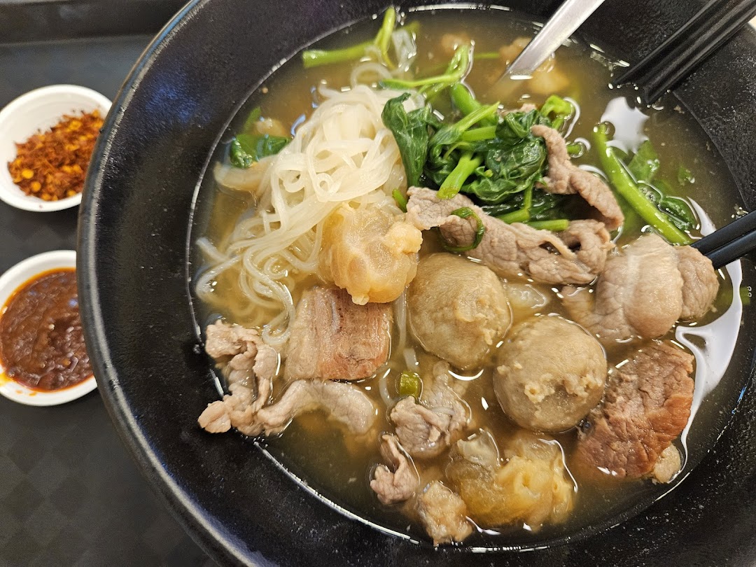 A close-up of a hearty bowl of Thai noodle soup with various meats, noodles, and greens, served with chili condiments.