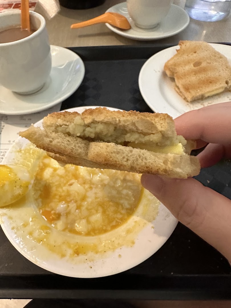 A hand holds kaya toast over a plate of soft-boiled eggs, with drinks and toast on a tray.