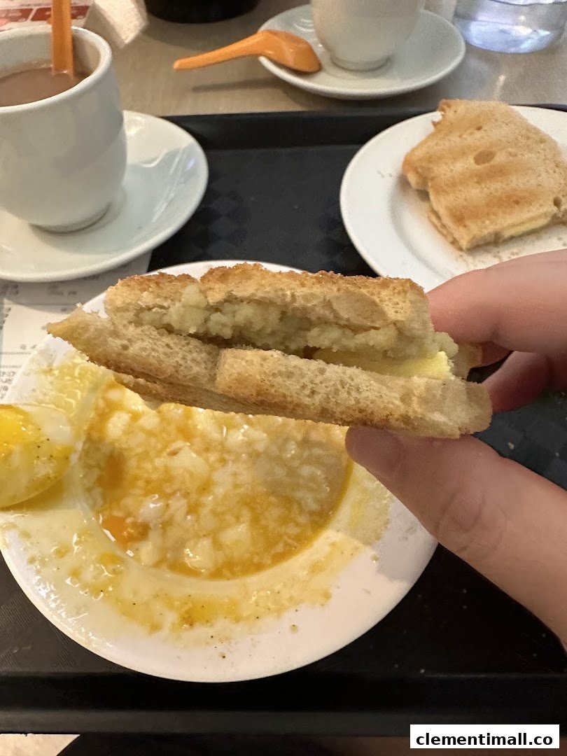 A hand holds kaya toast over a plate of soft-boiled eggs, with drinks and toast on a tray.