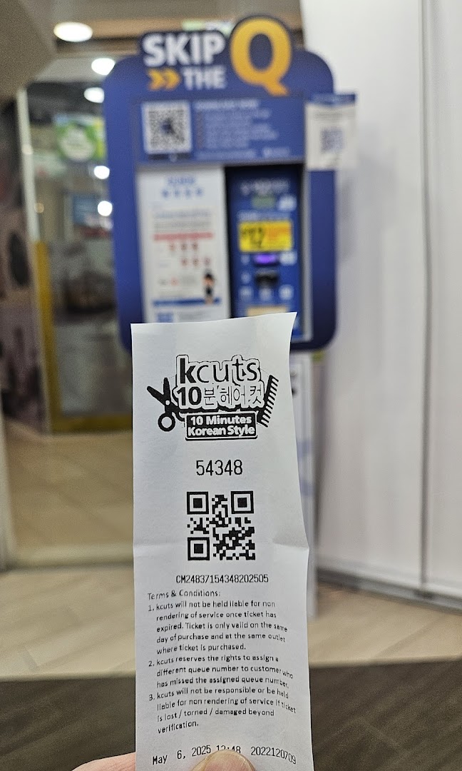 A customer holds a kcuts queue ticket in front of the 'SKIP THE Q' self-service machine.
