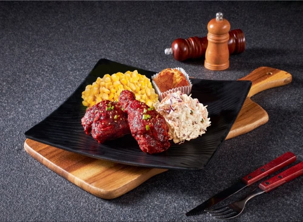 A delicious meal featuring red-glazed chicken, corn, coleslaw, and a muffin on a black plate.