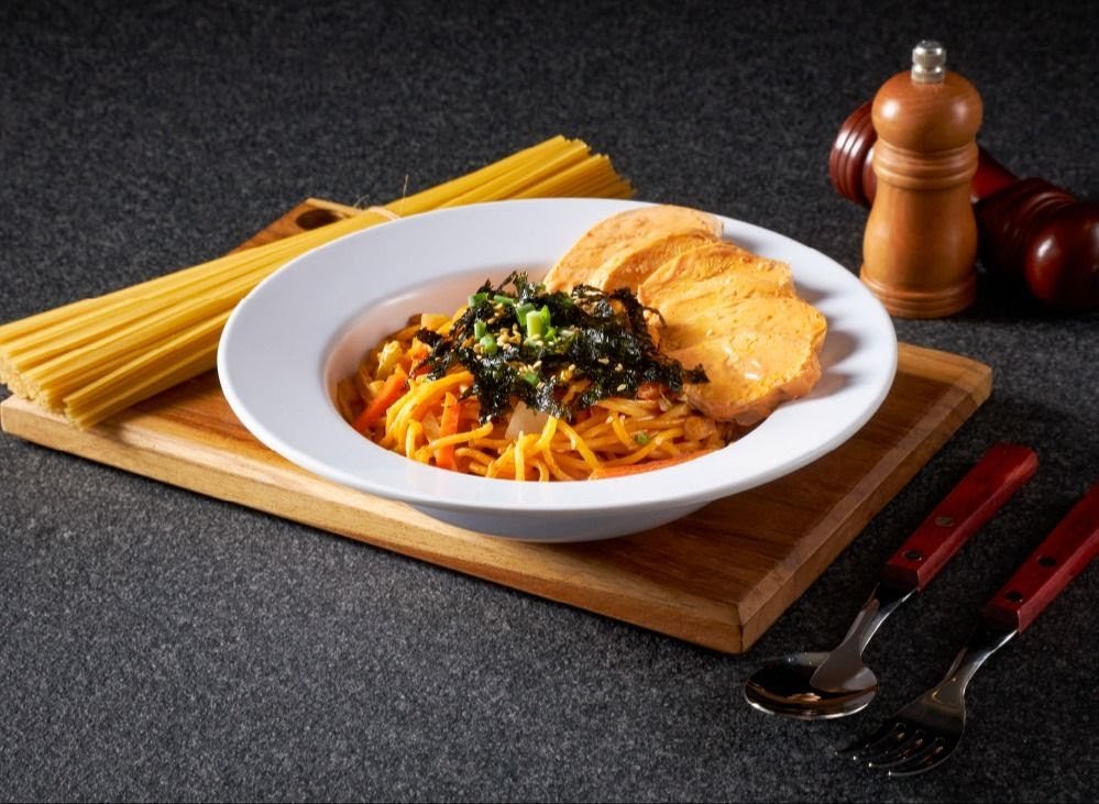 A vibrant pasta dish topped with seaweed and served with bread slices on a wooden board.